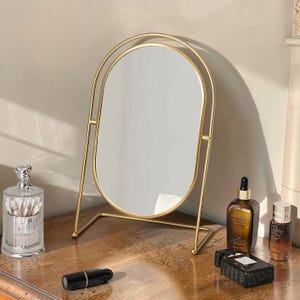 Puede incluir: Un espejo de mesa de metal dorado con un cristal ovalado suspendido dentro de un marco arqueado a juego. El espejo se apoya en una base sencilla y abierta, lo que permite una posición vertical estable sobre una superficie plana.