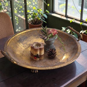 Peut inclure: Un grand plateau rond en métal doré avec deux poignées latérales et de petits pieds. À l'intérieur du plateau se trouvent une bougie cylindrique à la texture boisée, une petite urne grise en pierre avec des plantes succulentes et une pomme de pin.