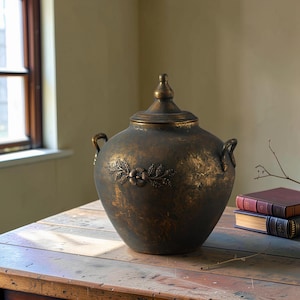 Op de afbeelding: Een donkerbronskleurige pot met deksel en een verweerde, gevlekte afwerking. De pot heeft twee gebogen handvatten aan de zijkanten en een centraal reliëf van een bloem omringd door bladeren. Het deksel loopt uit in een puntige vorm.