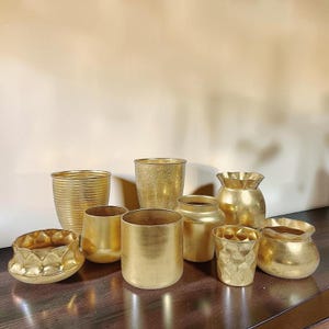 Puede incluir: Una colección de jarrones y recipientes decorativos de color dorado de varias formas y tamaños. Los recipientes incluyen diseños cilíndricos, redondeados y texturizados, todos con un acabado metálico. La disposición se muestra sobre una superficie de madera oscura.