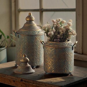 May include: Two cylindrical metal canisters with a blue and gold distressed finish, featuring an embossed geometric pattern, side handles, and small feet. One canister has a matching lid, while the other holds a bouquet of small white flowers.