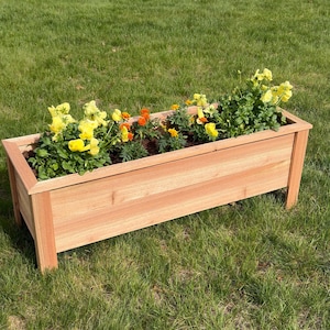 May include: A rectangular wooden planter box filled with yellow and orange flowers. The planter is made of light brown wood and sits on four short legs. The flowers are in full bloom, adding a splash of color against the green grass.