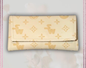 May include: Cream-colored wallet with a flap closure. The wallet features a repeating pattern of stylized "SW" and cross-shaped designs in a lighter cream color. The wallet is rectangular and appears to be made of a leather-like material.