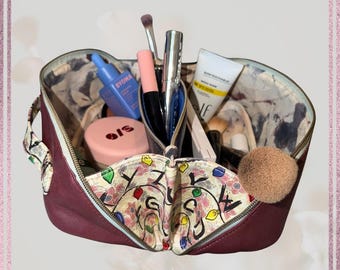 May include: A burgundy makeup bag with a floral and abstract patterned interior, filled with various cosmetic products. The bag is open, revealing brushes, tubes, and compacts. The bag has a zipper closure and a handle.