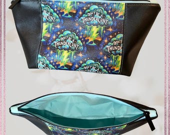 May include: Black cosmetic bag with a blue and green patterned design featuring the text "DON'T BE A THUNDERCUNT". The bag has a black zipper and a light blue interior. The bag is made of a durable material.