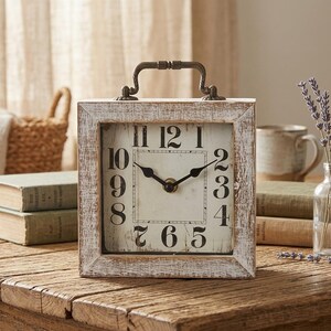 May include: A square analog clock with a distressed white wooden frame and a metal handle on top. The clock face is off-white with black Arabic numerals and black hour and minute hands. The design features a weathered finish on the frame.