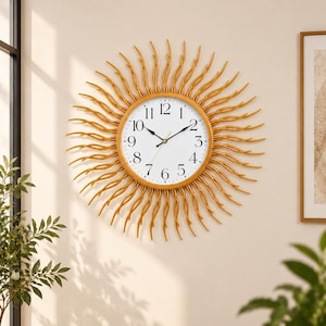 May include: A round wall clock with a gold-colored sunburst frame featuring radiating wavy metal spokes. The clock face is white with black Arabic numerals from one to twelve and two black hands indicating the time against a plain background.