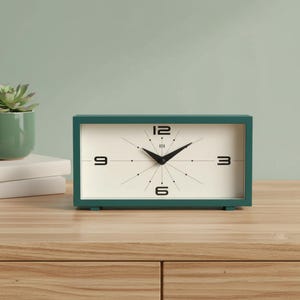May include: A rectangular table clock with a dark teal frame and an off-white face. The clock features black hour markers, thin radiating lines, and bold black numerals at the 3, 6, 9, and 12 o'clock positions. Two black hands indicate the time.