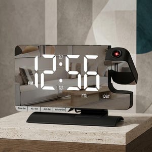 May include: A rectangular digital alarm clock with a reflective mirror finish display showing the time 12:56 PM, the day FRI, and DST. It features a black base, a side projection arm with a red lens, and buttons labeled Time-Set, AL1-Set, AL2-Set, and Volume/Dst.