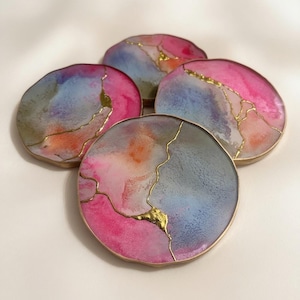 May include: A set of four circular coasters featuring a marbled blend of pink, blue, and orange colors. Each coaster has a smooth, glossy finish with metallic gold-colored veins running across the surface and a thin gold-colored metal rim.