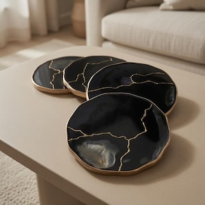May include: A set of four round coasters with a glossy black finish, featuring irregular gold-colored vein patterns that mimic natural stone. Each coaster has a smooth, slightly uneven edge and a reflective surface with subtle gold accents.