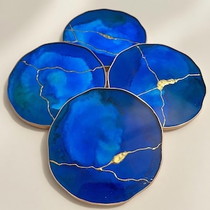 May include: A set of four circular coasters featuring a deep blue and teal marbled resin finish. Each coaster is accented with metallic gold veins that mimic the appearance of kintsugi, creating a distinct pattern across the smooth, glossy surfaces.