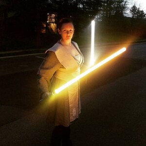 Puede incluir: Una persona vestida con una t&uacute;nica marr&oacute;n claro sostiene dos sables de luz brillantes, uno amarillo y otro blanco, en un entorno oscuro.