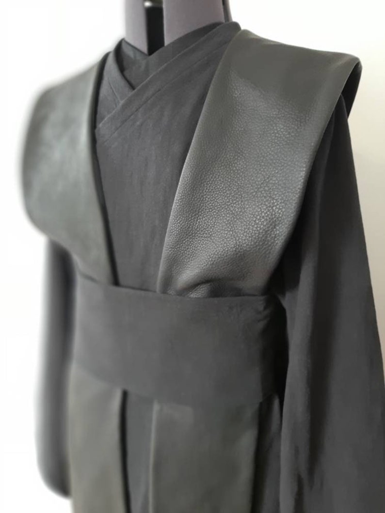 Puede incluir: T&uacute;nica negra con cuello de cuero y cintur&oacute;n de tela negro. La t&uacute;nica tiene manga larga y cuello en V.