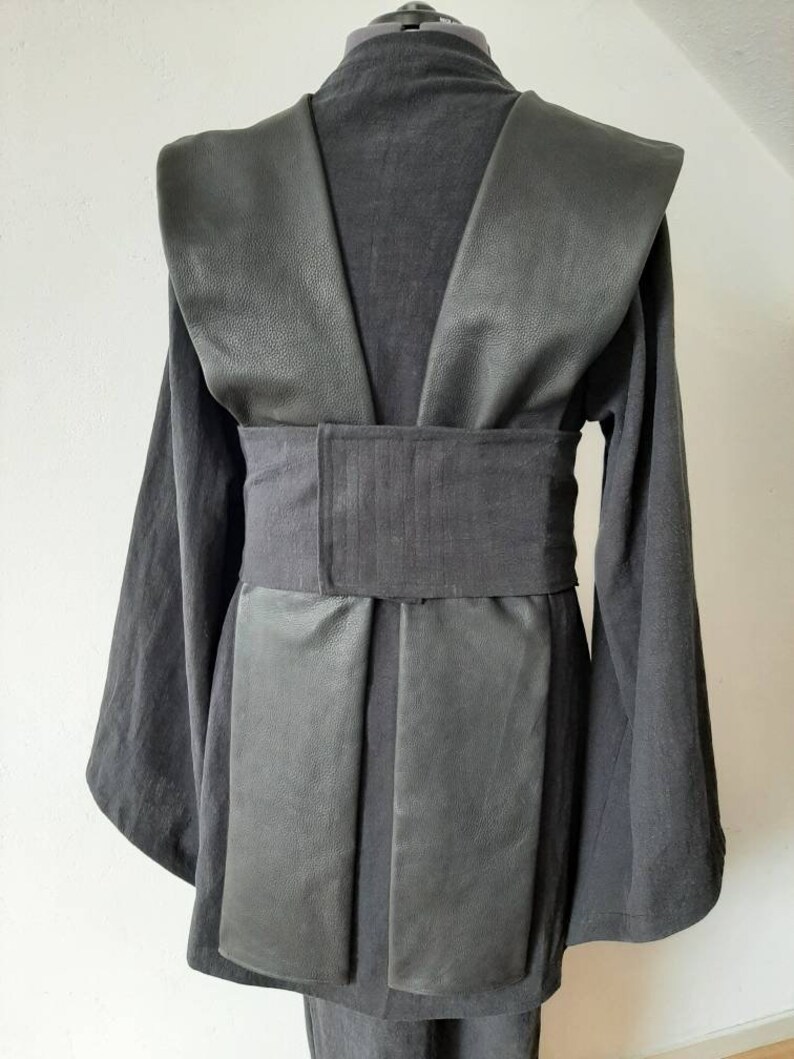 Puede incluir: T&uacute;nica negra con cuello de cuero y cintur&oacute;n de tela negro. La t&uacute;nica tiene mangas largas y cuello en V.