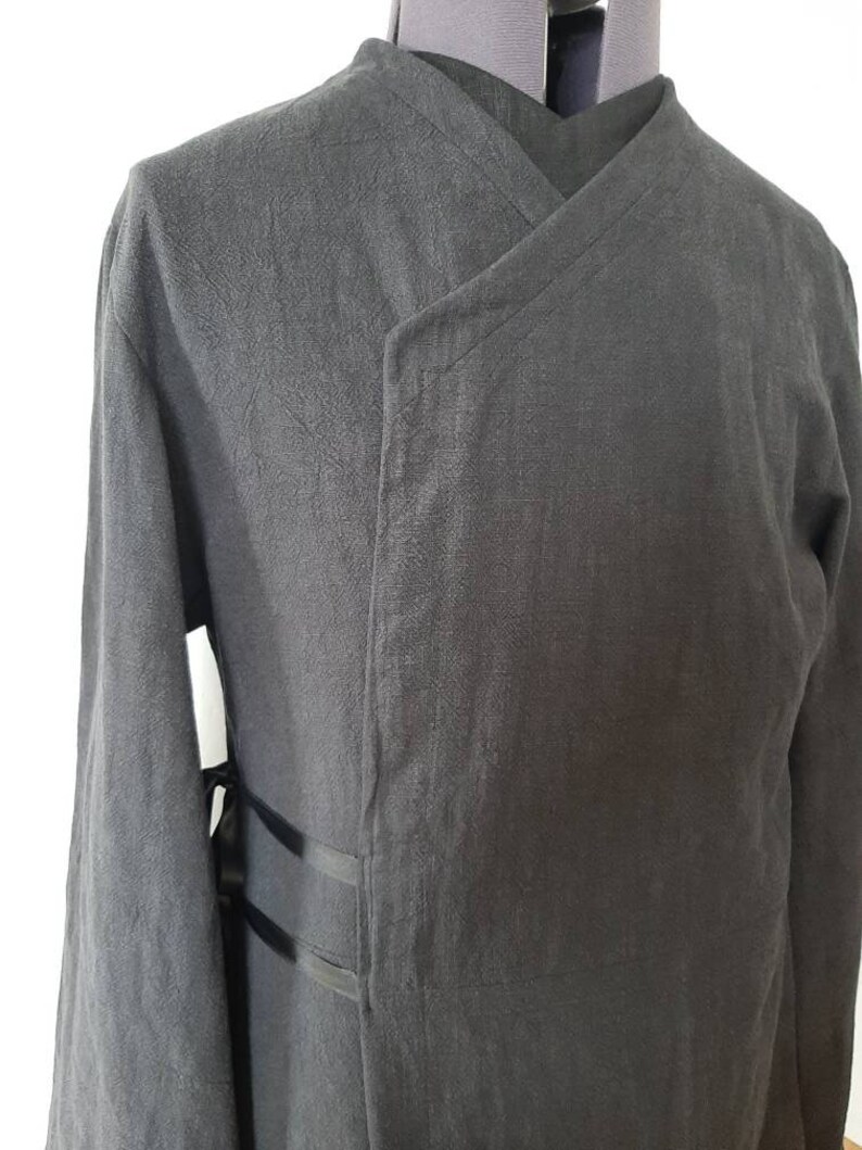 Puede incluir: Una chaqueta negra de manga larga con cuello mandar&iacute;n y cierre de envoltura. La chaqueta tiene una tela texturizada y dos correas negras en el lateral.
