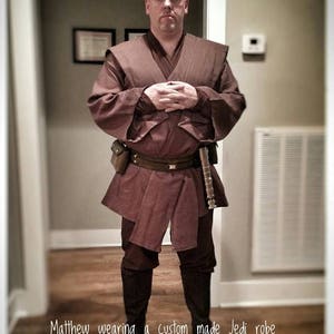 Puede incluir: Una persona vistiendo una t&uacute;nica Jedi marr&oacute;n con un cintur&oacute;n marr&oacute;n y un sable de luz. El texto "Matthew wearing a custom made Jedi robe BY ETSY.COM/SHOP/AVALONDESIGNSNL" est&aacute; en la parte inferior de la imagen.