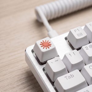 Op de afbeelding: Close-up van een wit mechanisch toetsenbord met een zilveren frame. Het toetsenbord heeft witte keycaps met een rood sunburst-ontwerp op één toets. Een witte spiraalkabel is aangesloten op het toetsenbord.