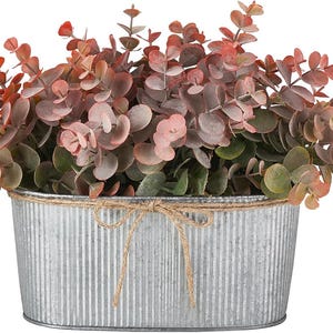 May include: Oval galvanized metal planter filled with faux eucalyptus. The leaves are a mix of green, pink, and red. A natural twine bow adorns the front of the planter, adding a rustic touch. Perfect for home decor.