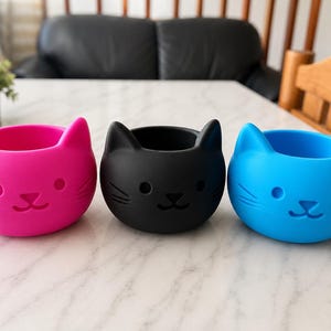 Puede incluir: Tres macetas con forma de gato en colores brillantes: rosa intenso, negro y azul. Cada maceta tiene un cuerpo redondo con un diseño de cara de gato, que incluye orejas, ojos y nariz. Las macetas están hechas de un material liso y están sobre una superficie blanca.