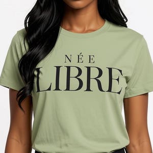 May include: A sage green short-sleeved t-shirt featuring the text NÉE LIBRE printed in black across the chest. The shirt has a crew neckline and a standard fit, paired with black trousers. The design is centered on the front of the garment.