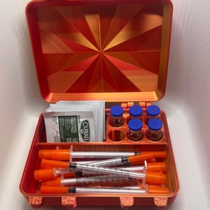 Könnte beinhalten: Ein offener orangefarbener Koffer mit einem geometrischen Design auf dem Deckel. Im Inneren befinden sich Fläschchen mit blauen Kappen, Spritzen und Alkoholtupfern. Der Koffer dient zur Organisation und Aufbewahrung von medizinischen Utensilien.