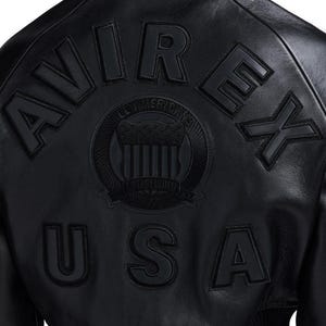 May include: A black leather jacket featuring the embossed text AVIREX USA in large capital letters. Centered between the text is a circular emblem with the words ALL AMERICAN and ESTABLISHED N.Y. surrounding a shield graphic design.