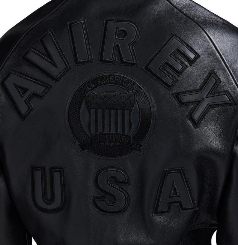 May include: A black leather jacket featuring the embossed text AVIREX USA in large capital letters. Centered between the text is a circular emblem with the words ALL AMERICAN and ESTABLISHED N.Y. surrounding a shield graphic design.