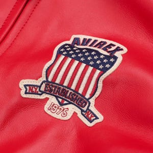 May include: A bright red leather jacket featuring an embroidered patch on the chest. The patch displays a shield with red and white stripes and blue stars, the text AVIREX, ESTABLISHED 1975, and N.Y. on a cream-colored background with blue stitching.