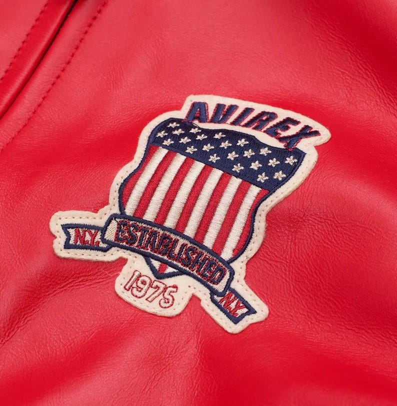 May include: A bright red leather jacket featuring an embroidered patch on the chest. The patch displays a shield with red and white stripes and blue stars, the text AVIREX, ESTABLISHED 1975, and N.Y. on a cream-colored background with blue stitching.