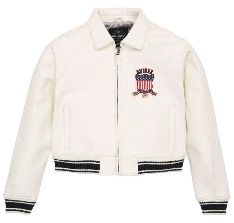May include: An off-white bomber jacket with a classic collar, a full-length front zipper, and long sleeves. The chest features an embroidered patch with the text AVIREX and a flag design. The hem and cuffs have black and white striped ribbed trim.