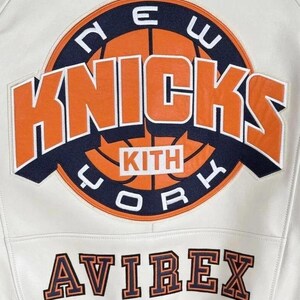 Op de afbeelding: Een wit leren jack met een grote ronde patch op de achterkant met een basketbalafbeelding, de tekst NEW YORK KNICKS en het woord KITH in een centraal kader. Onder de patch is de merknaam AVIREX geborduurd in oranje en marineblauw.