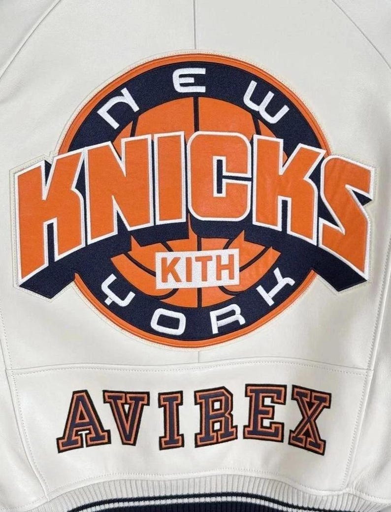 Op de afbeelding: Een wit leren jack met een grote ronde patch op de achterkant met een basketbalafbeelding, de tekst NEW YORK KNICKS en het woord KITH in een centraal kader. Onder de patch is de merknaam AVIREX geborduurd in oranje en marineblauw.