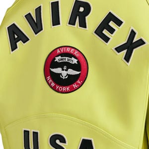 May include: A bright lime green jacket featuring the black text AVIREX arched across the upper back. Below the text is a circular patch with a red border, black center, and white graphic of wings and an anchor, with the text AVIREX SINCE 1975 NEW YORK N.Y.