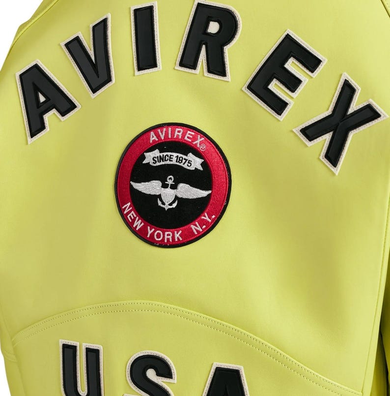 May include: A bright lime green jacket featuring the black text AVIREX arched across the upper back. Below the text is a circular patch with a red border, black center, and white graphic of wings and an anchor, with the text AVIREX SINCE 1975 NEW YORK N.Y.