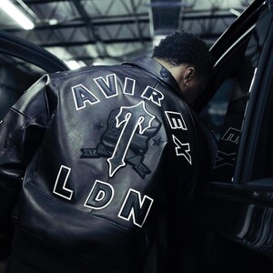 May include: A black leather jacket featuring the text AVIREX in an arc across the upper back, with the letters LDN positioned below. A large central emblem displays a stylized letter T with a shield design, surrounded by small star accents.