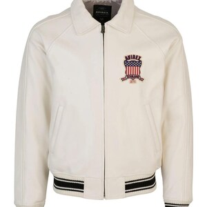 Puede incluir: Una chaqueta color blanco hueso con cremallera frontal completa, cuello cl&aacute;sico y pu&ntilde;os y dobladillo acanalados con rayas negras y blancas. Un parche en el pecho izquierdo muestra el texto AVIREX sobre un escudo con estrellas y rayas y el a&ntilde;o 1975.