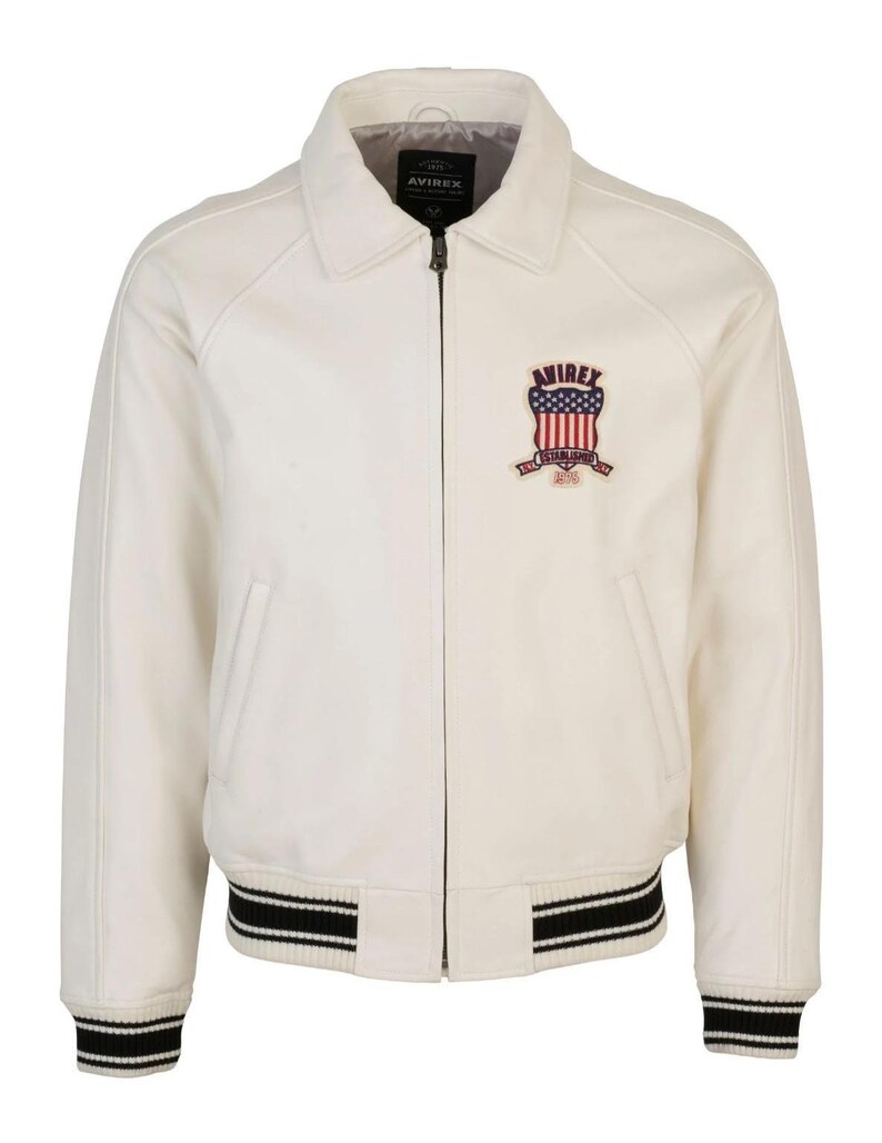 Puede incluir: Una chaqueta color blanco hueso con cremallera frontal completa, cuello cl&aacute;sico y pu&ntilde;os y dobladillo acanalados con rayas negras y blancas. Un parche en el pecho izquierdo muestra el texto AVIREX sobre un escudo con estrellas y rayas y el a&ntilde;o 1975.
