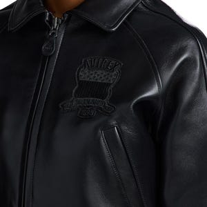 May include: A black leather jacket featuring a full-length front zipper with a branded pull tab, a classic collar, and a black embroidered patch on the chest. The patch displays the text "AVIREX" above a graphic and "ESTABLISHED 1975".