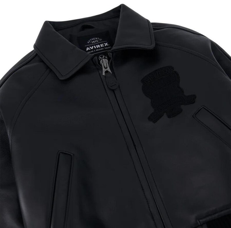 May include: A black leather jacket featuring a classic collar, a full-length front zipper with a branded pull, and two side pockets. An embroidered patch is visible on the chest, and the interior label displays the text AVIREX AUTHENTIC 1975.