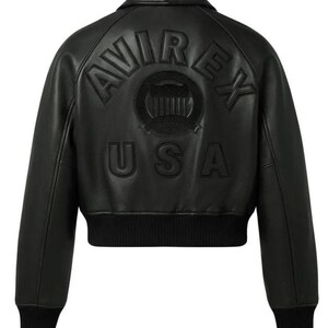 May include: A black leather bomber jacket featuring a ribbed collar, cuffs, and hem. The back displays embossed lettering that reads AVIREX USA, with a circular emblem positioned between the words, all rendered in a matching black tone.