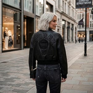 May include: A black leather bomber jacket featuring the embossed text AVIREX USA on the back. The jacket has a ribbed hem and cuffs, paired with dark grey denim jeans. A black shoulder bag with a gold-toned metal strap attachment is visible.
