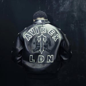 May include: A black leather jacket featuring a large white embroidered logo on the back. The text AVIREX is arched above a central T-shaped emblem, with the letters LDN positioned below. The jacket has a classic collar and ribbed hem detailing.