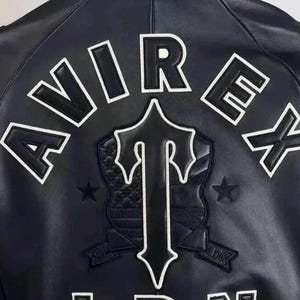 May include: A dark navy blue leather jacket featuring the white embroidered text AVIREX and LDN on the back. A large central black and white shield emblem with a stylized letter T is positioned between the text, flanked by two small stars.