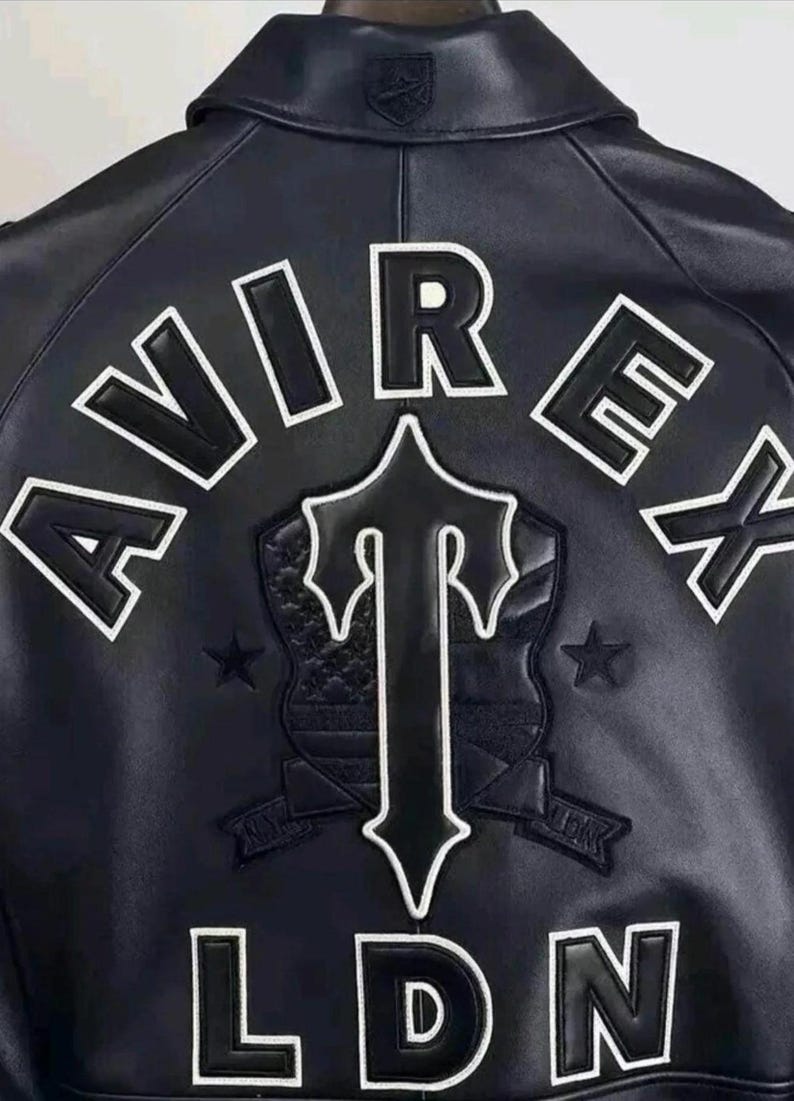 May include: A dark navy blue leather jacket featuring the white embroidered text AVIREX and LDN on the back. A large central black and white shield emblem with a stylized letter T is positioned between the text, flanked by two small stars.