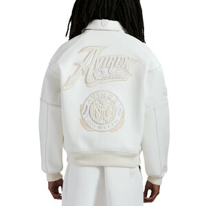 May include: An off-white varsity jacket featuring a large embroidered Avirex New York logo on the upper back and a circular Avirex Trade Mark NYC patch below it. The jacket has a ribbed collar, cuffs, and hem, with perforated leather sleeve panels.