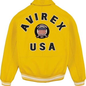 Pu&ograve; includere: Una giacca varsity giallo brillante con la scritta AVIREX in lettere nere arcuata sopra una toppa circolare con uno scudo a bandiera americana rosso, bianco e blu e il testo ALL AMERICANS ESTABLISHED 1975, con la scritta USA in nero sotto.