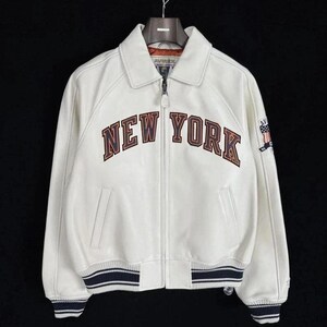 Op de afbeelding: Een wit leren jack met een ritssluiting aan de voorzijde, een klassieke kraag en geribbelde boorden en zoom met marineblauwe en witte strepen. Op de borst staat de tekst NEW YORK in donkerbruine letters en een vlagpatch op de mouw.