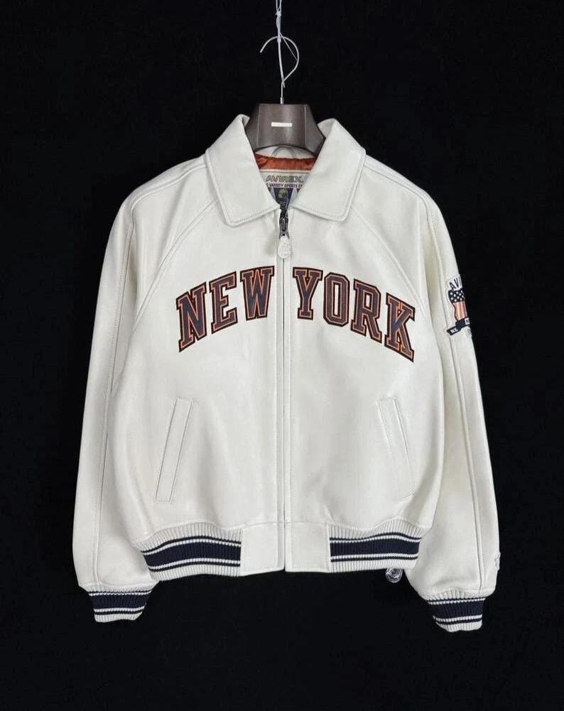 Op de afbeelding: Een wit leren jack met een ritssluiting aan de voorzijde, een klassieke kraag en geribbelde boorden en zoom met marineblauwe en witte strepen. Op de borst staat de tekst NEW YORK in donkerbruine letters en een vlagpatch op de mouw.