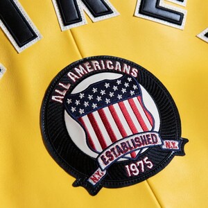 Pu&ograve; includere: Una giacca giallo brillante con una grande toppa circolare ricamata raffigurante uno scudo con strisce rosse e bianche e stelle blu. Il testo recita ALL AMERICANS ESTABLISHED 1975 N.Y. In alto sono visibili grandi lettere a blocchi bianche e nere.